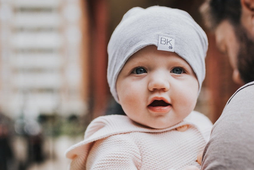 Baby in a light pink outfit and gray beanie being held by an adult outdoors.
